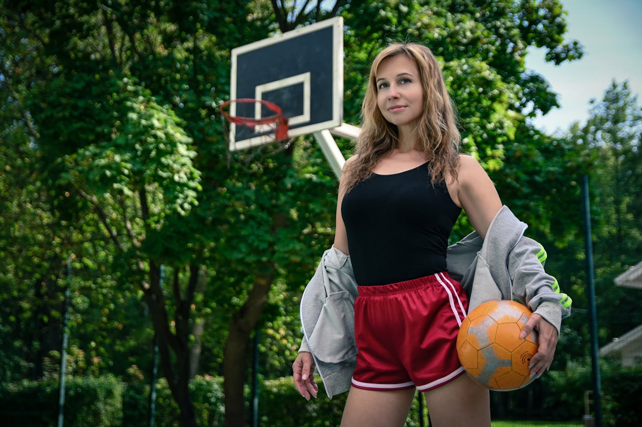 Confident woman posing with a basketball on an outdoor court surrounded by lush greenery.